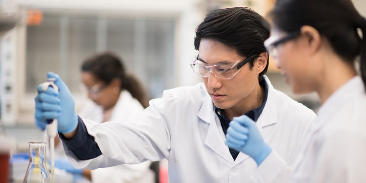 A team of male and female Biochemists, use pipettes to fill samples vials for testing. They are seated at a desk filed with other instruments, flasks and sharps bins. They are all wearing protective eye wear, medical gloves and lab coats for protection.