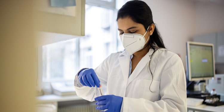 Woman standing in the medical laboratory looking at blood samples. Female researcher wearing protective face mask and gloves handling samples in lab.